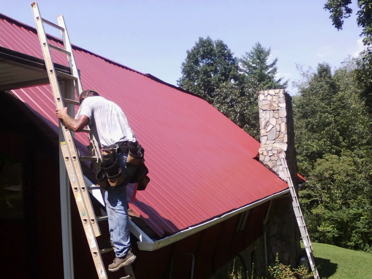 Roofer on a ladder beside a red metal roof and stone chimney — Soffit & Fascia Repair by Roofing Brothers in Maryland Line
